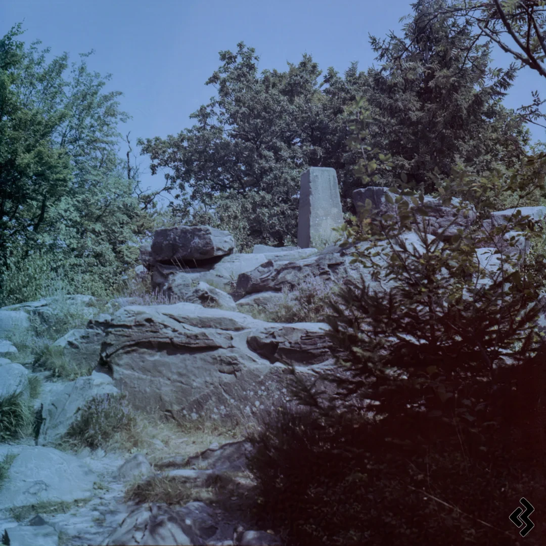Photo of large rocks with a stone obelisk on top marking a mountain top. The weather is sunny with a cloudless sky, in the background are some trees and bushes