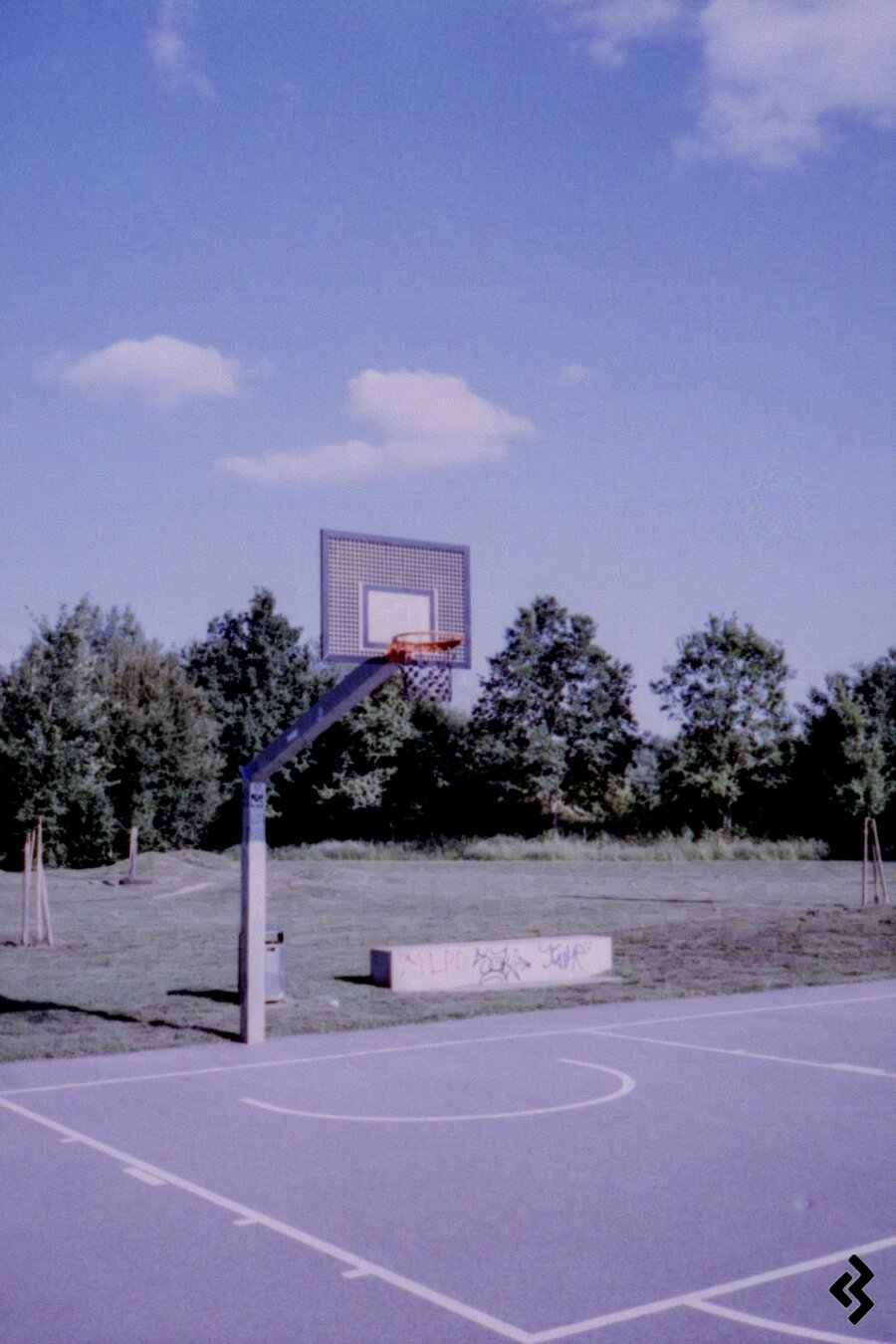 Photo of a basketball court in a park. The surface of the court is asphalt, and the basketball hoop is made of steel. In the background, a closely mowed lawn and a few trees can be seen. At the edge of the basketball court, directly behind the hoop, is a minimalist concrete bench covered in graffiti. The sky is blue with scattered white clouds.