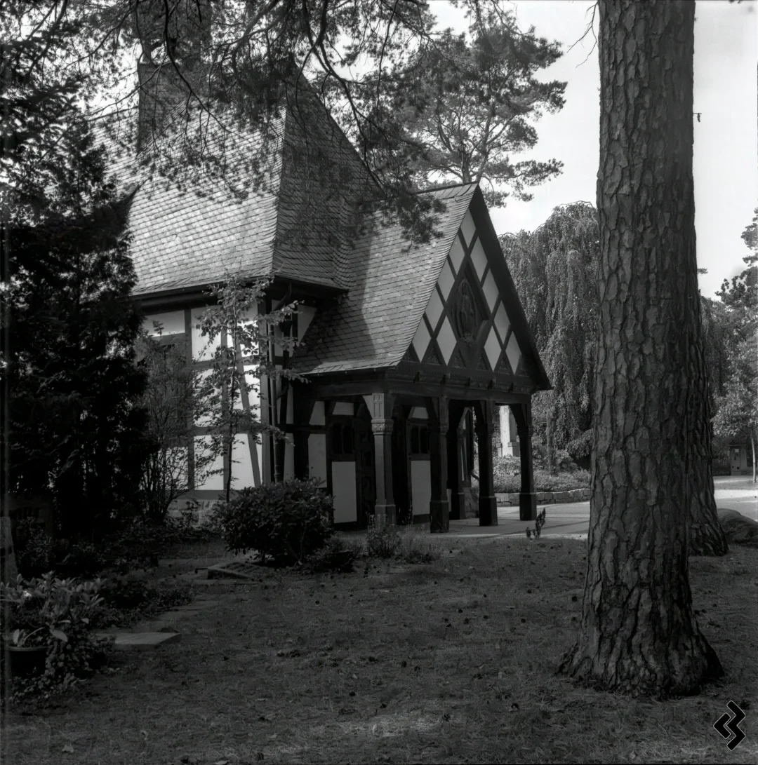 A black and white photo of a small, quaint chapel with a steeply pitched roof and timber frame architecture. The building features decorative timber framing and a covered porch supported by wooden columns. It is surrounded by tall pine trees, bushes, and a grassy yard with scattered pine cones.