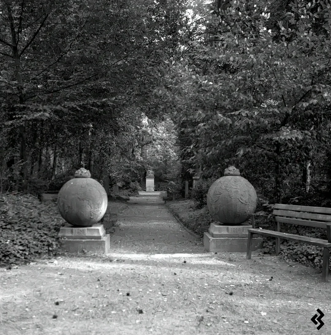 Square format black and white photography of a park. Two stone globes stand on the left and right at the beginning of a path. The path is lined with trees, and at the end is a clearing with a statue. Next to the right-hand globe is a park bench.