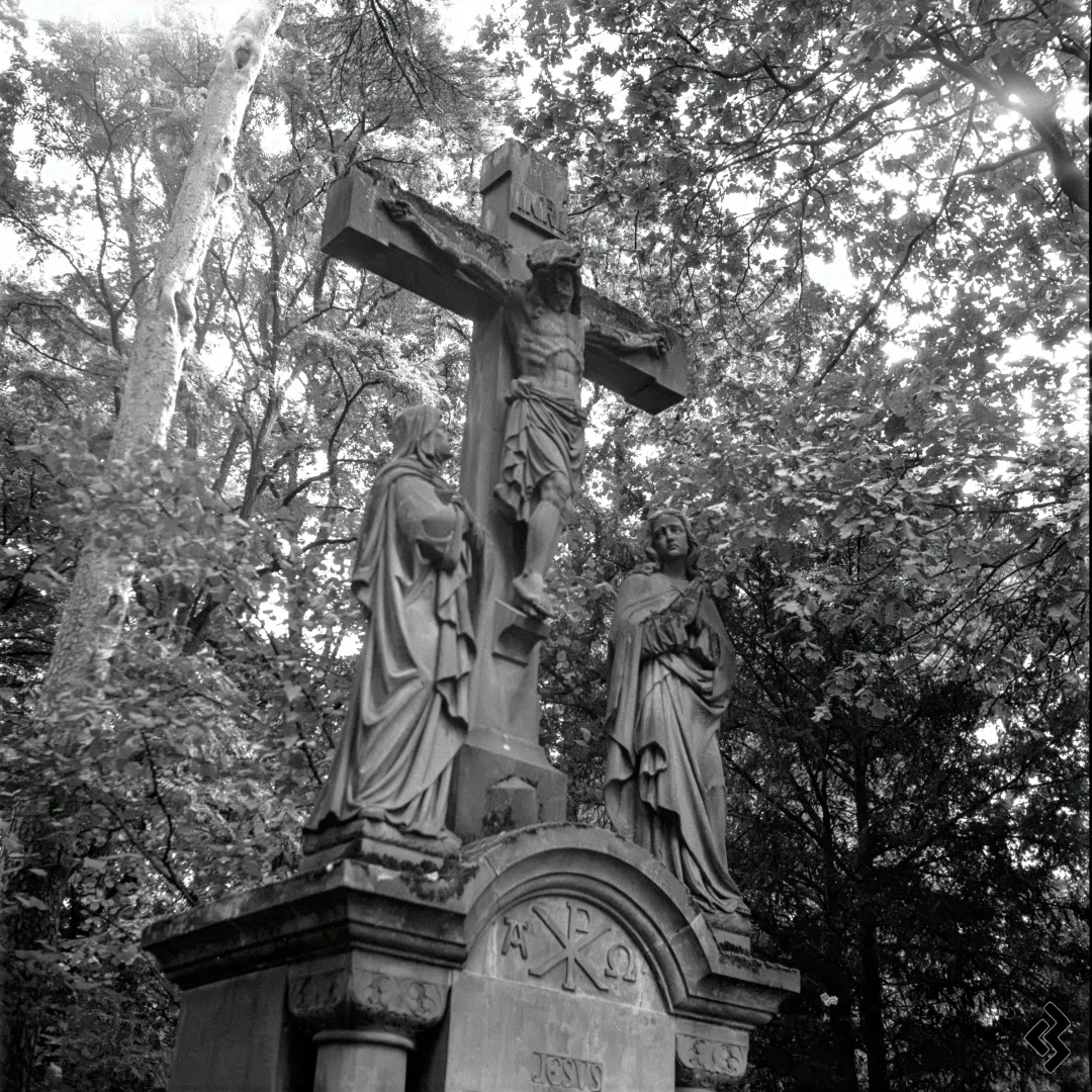 Square format black and white photo of a cross at a cemetery. Two statues of women are placed below the cross, a little bit of moss is growing on the statues. The whole statue group is located in a forest area, with sunlight breaking through the leaves in the background.