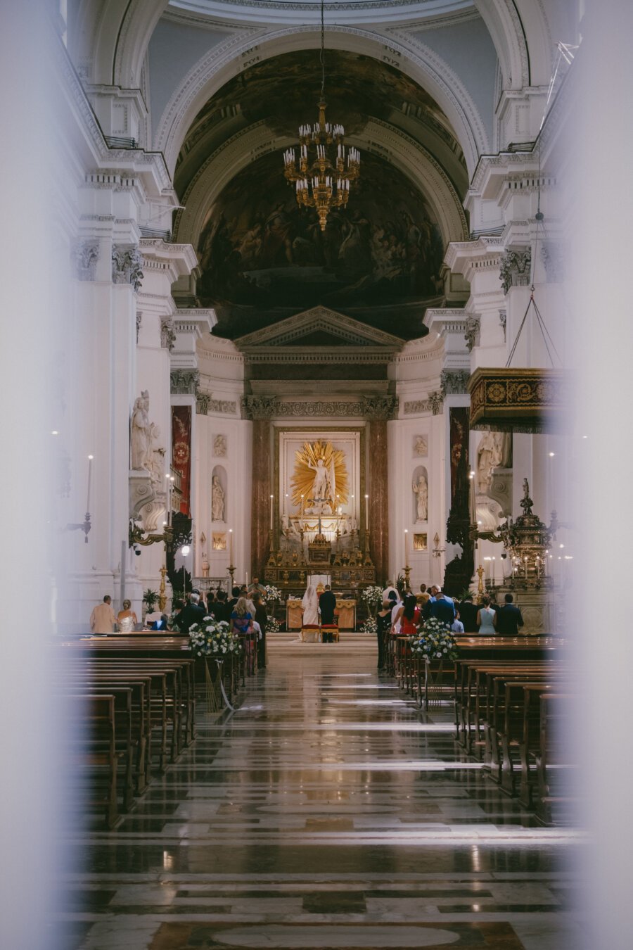 ein kirchgang an dessen ende der altar ist, wo gerade ein paar getraut wird durch den zaun vor der kirche, deren türe weit offen steht, fotografiert