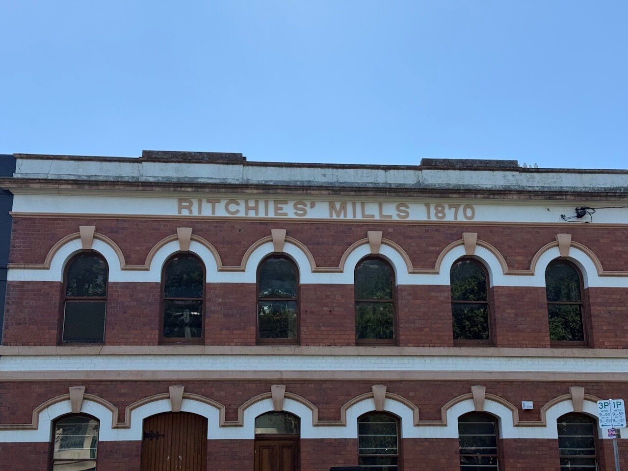 Large gold signage says “Richie’s Mills 1870”. The signage sits above two rows of regularly spaced black windows with cream architraves that hop across the top of each window to form a wave across the facade.