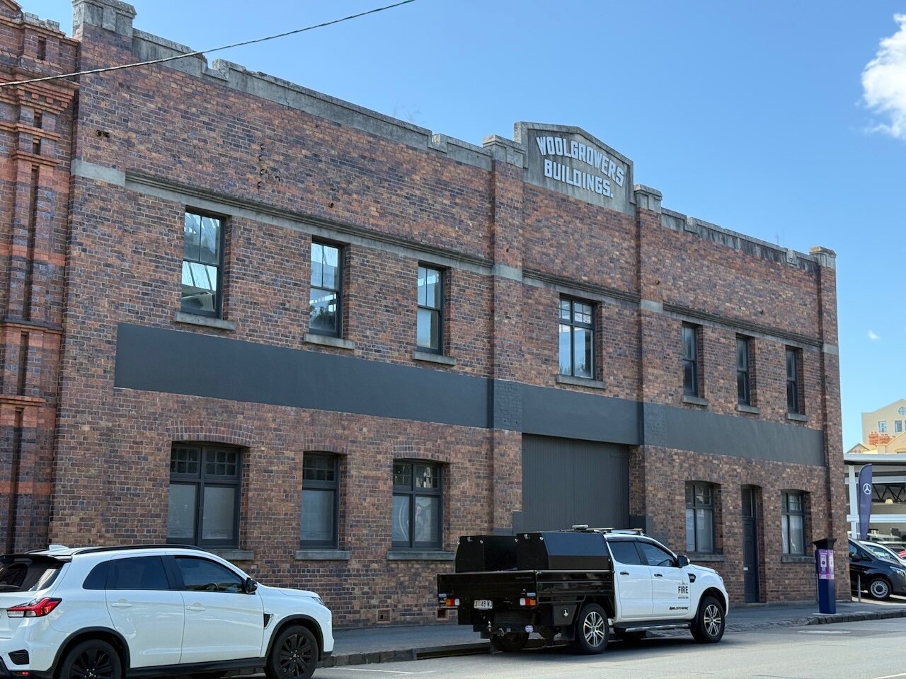 A brick built building with regularly spaced black windows on two levels is split in half by a roll up garage door. The signage says “Woolgrowers buildings”. Modern cars sit on the road in front and juxtaposed the older style building design.