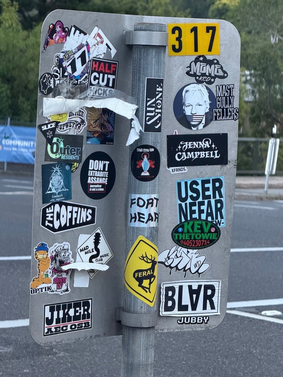 Waiting at a traffic light, looking at the back of a street sign intended for drivers on the opposite side of the street. The back is covered in stickers of varying sizes and colours. One says “Don’t extradite Assange; Journalism is not a crime”, another is a photo of Assange gagged by a USA flag. Others appear to be band names like “Half Cut Hicks”, “Mast Gully Fellas” and “Colem Dance Club”. There are many others, including one for “Kev the Towie” and another that just says “Jubby”.