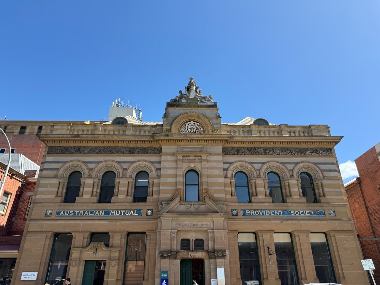 A stone-built building with blue and gold mosaic panels saying “Australia Mutual Provident Society”. The stone above the door dates the building to 1849.