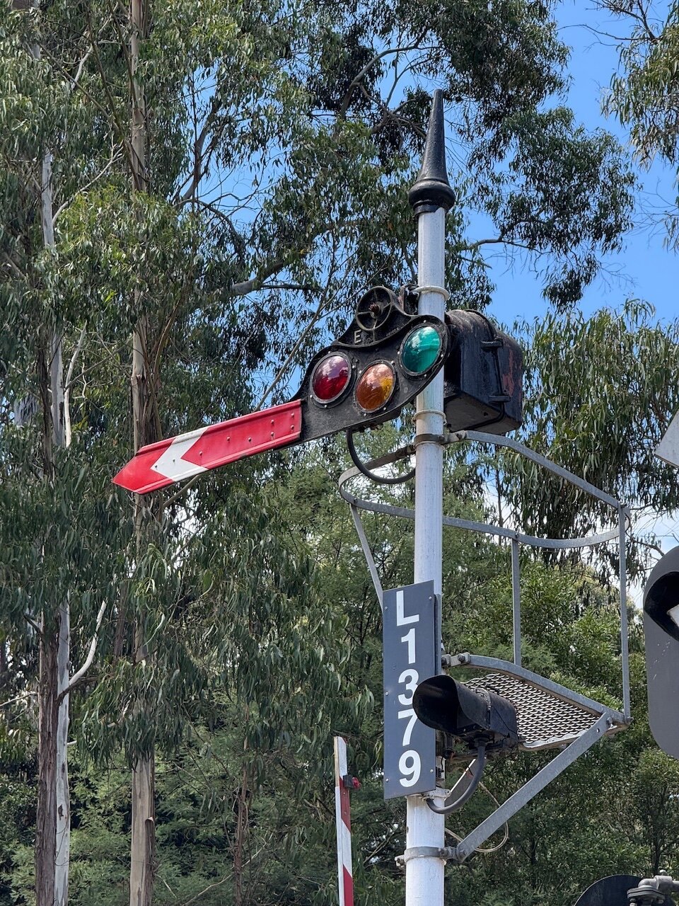An old train signal with red, yellow and green lenses attached to a red tail with a white arrow. The signal is labelled “L1379”. In the background there are Eucalyptus trees.