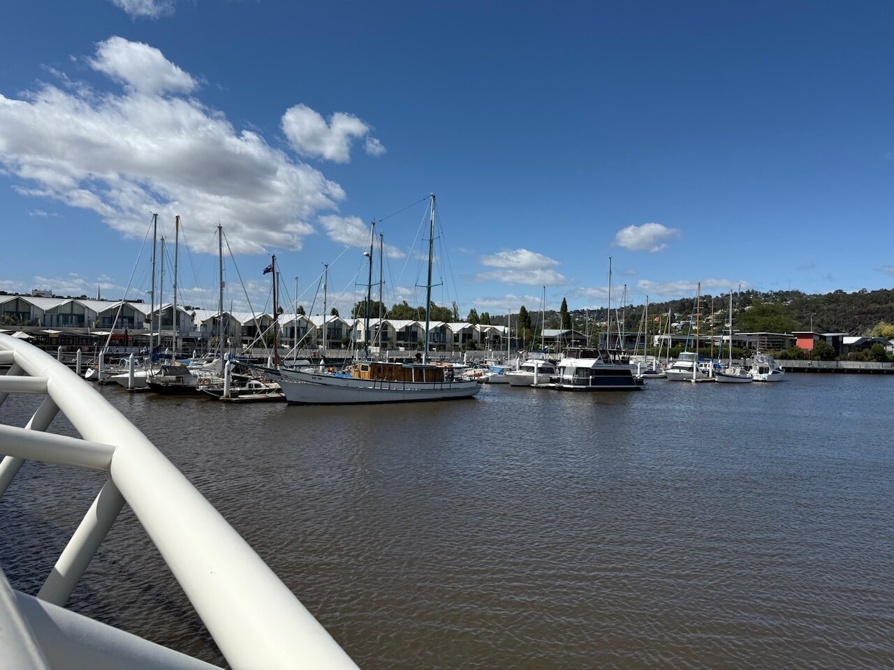 A small marina filled with boats viewed from a modern white bridge. There are larger sail-based boats at the front with smaller motorboats tucked behind. The tide is high and there are stout houses overlooking the back in the background.