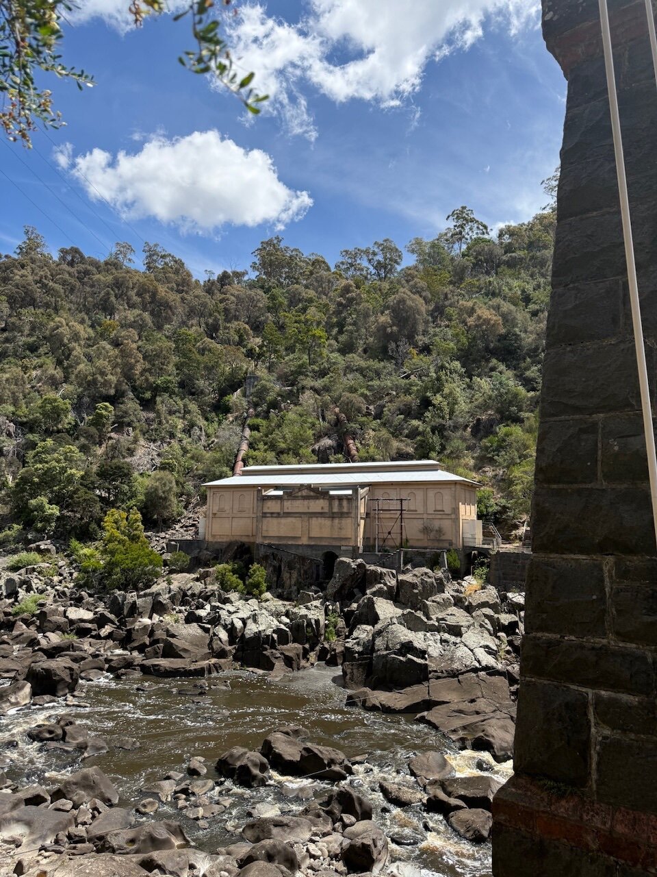 A look back across the South Esk River canyon, past the bridge archway, onto the disused Duck Reach Hydroelectric Power Station building. It’s painted a beige colour and the old pipework delivering water into the building can be seen on the hill behind. The banks of the Esk are rocky and the river water level is low.