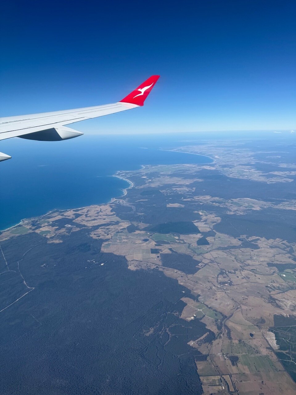A plane wing, the tip is red and has a kangaroo logo (the logo of Qantas Airlines). Below the aircraft is a landmass (Tasmania) and you can see fields and the sea lapping against the beaches below. The skies are clear and you can see all the way to the horizon.