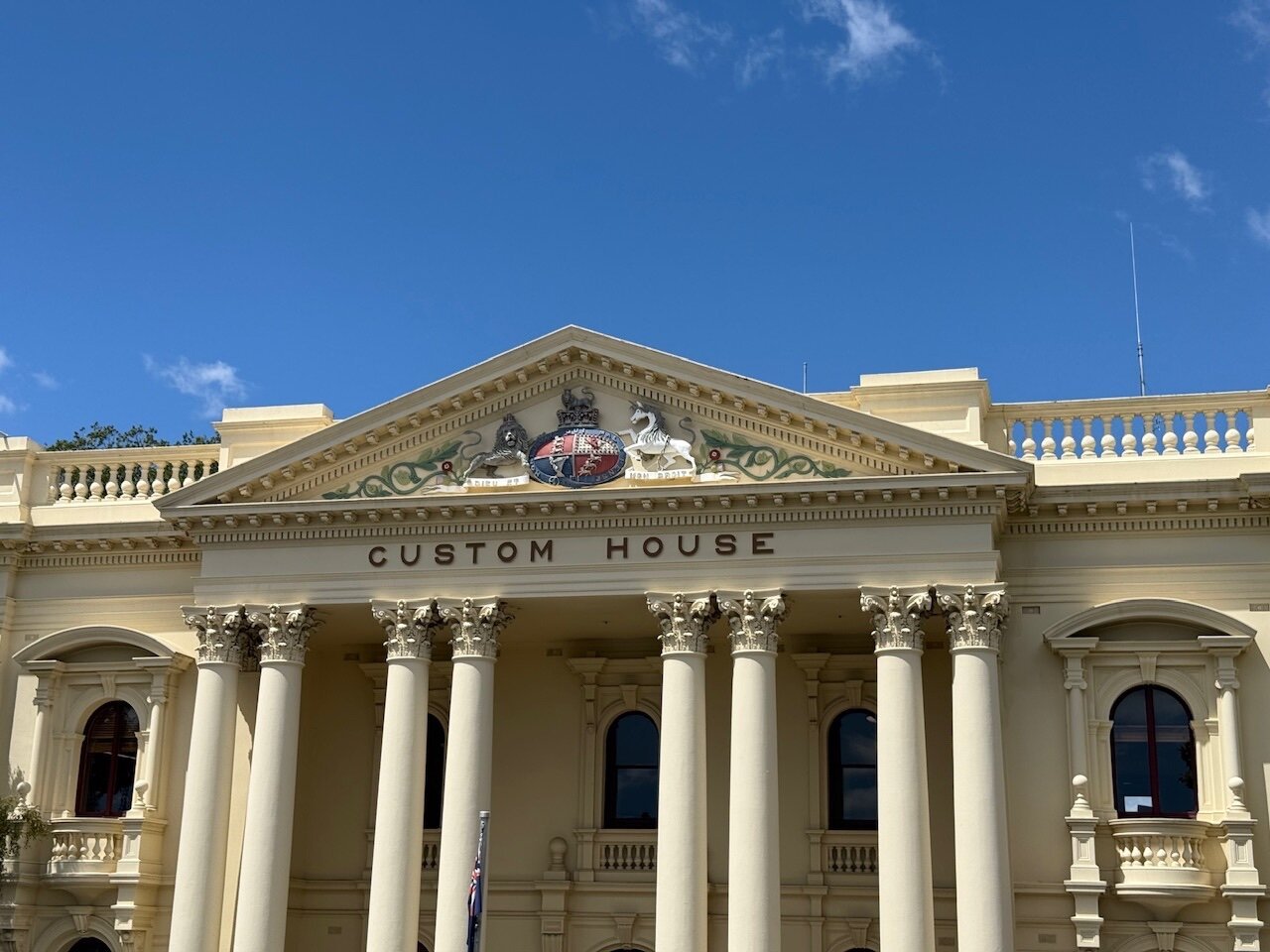 A columned building built in cream marble is topped with a triangular roof. The triangular shape contains a U.K. seal with the usual Lion and Unicorn symbology and “Dieu et mon droit” mantra. Large black letters spell out “Custom House” below. A flagpole with the Australian flag stands in front of the building.