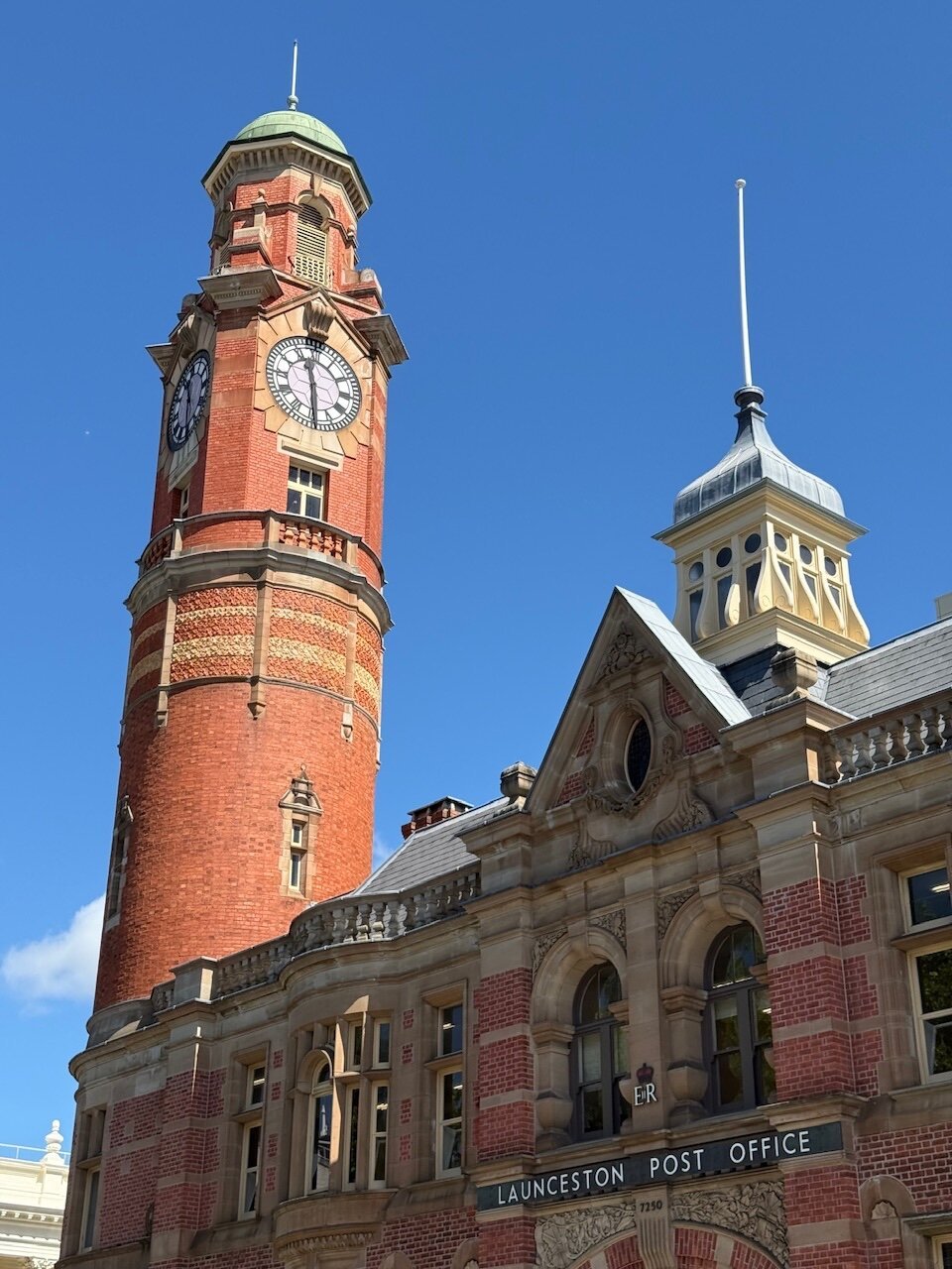 A brick-built building labelled “Launceston Post Office”. A circular clock tower stands proudly into the sunshine while the main building sits in shadow. The sky is deep blue in the background and the time displayed on the clock face is 12:30