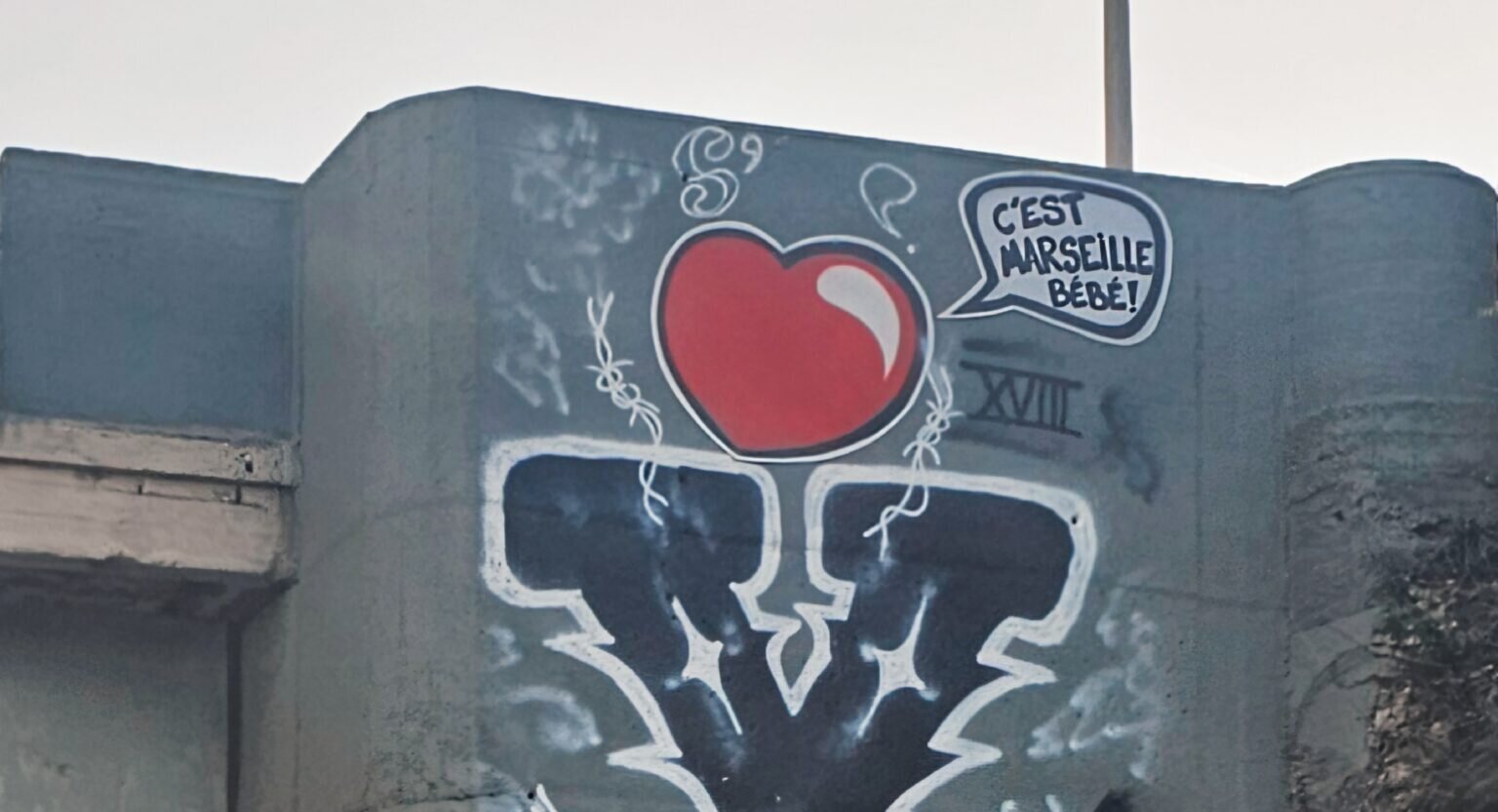 détail d'une photo (celle du billet de blog) : sur un pilone d'un ouvrage d'art, un gros coeur rouge assorti d'un phylactère dans lequel est écrit : C'EST MARSEILLE BEBE !