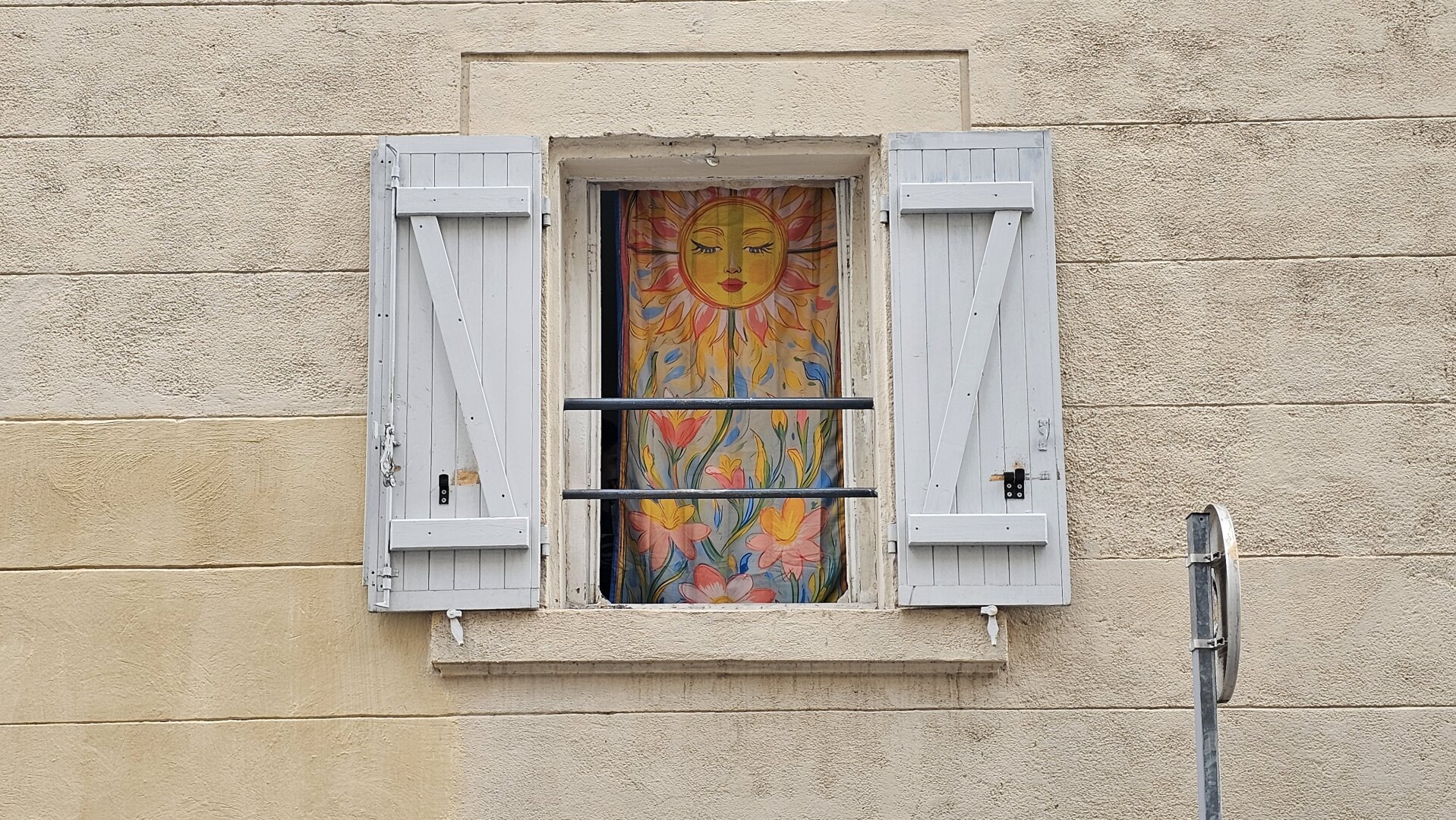 une fenêtre, volets ouverts, un rideau colorée représentant un soleil et des fleurs