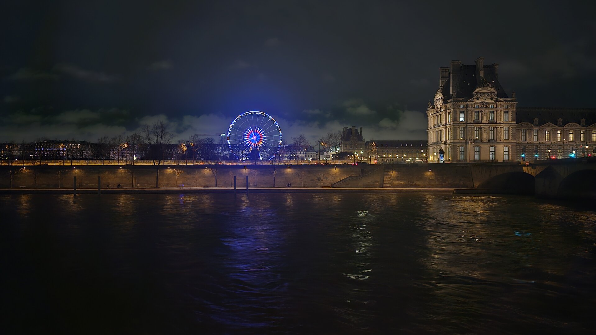 Photo nocturne. Au premier plan la Seine, au second plan la grand roue au Jardin des Tuileries puis le Louvre à droite
