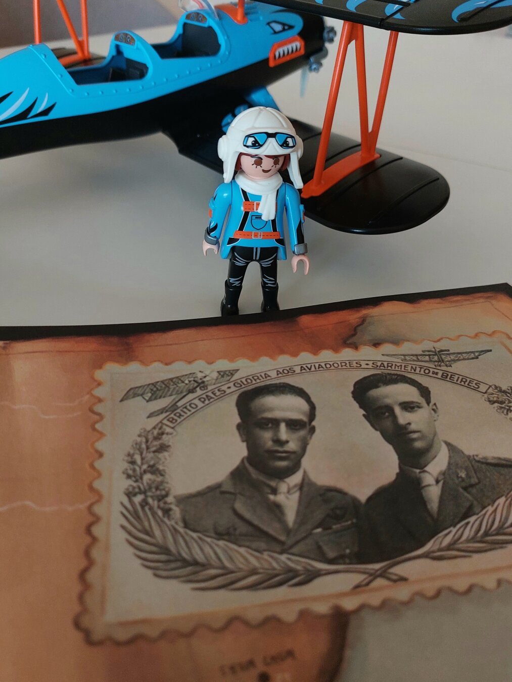 Piloto menina playmobil com capacete branco e roupa azul a olhar para a ilustração do livro onde aparecem os pilotos Brito Pais e Sarmento de Beires. Ao fundo o seu avião bimotor azul