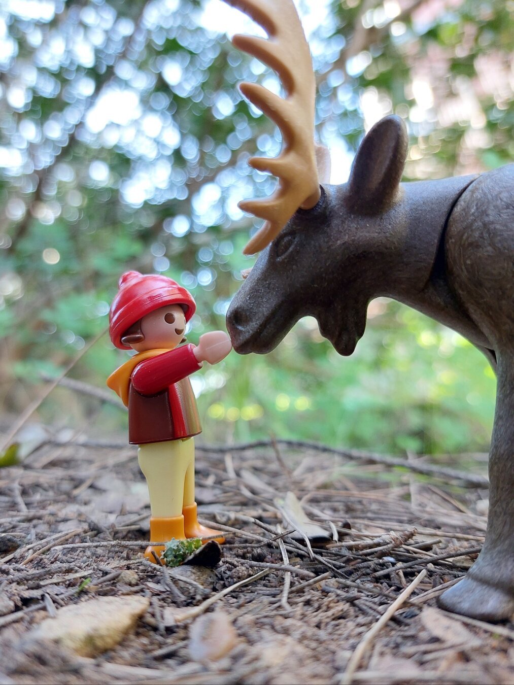 Criança playmobil vestida de vermelho, calças amarelas, acariciando um alce na floresta