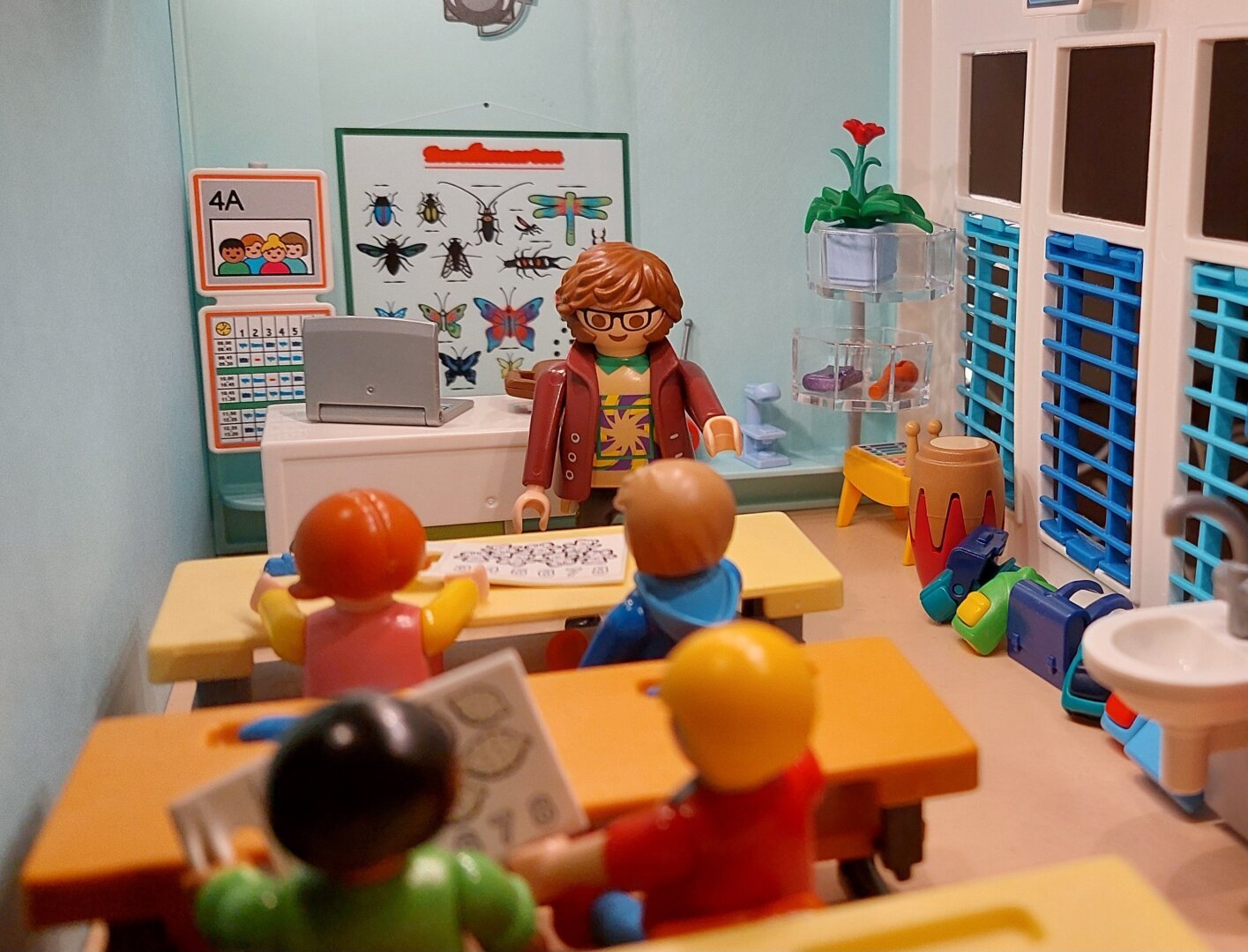 Varias crianças playmobil, dentro de uma sala de aula playmobil de paredes azuis, mesas aparelas, professor de roupa e cabelo castanho, óculos