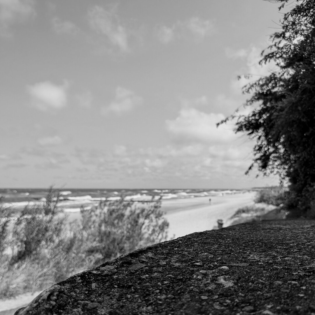 black and white photo of the sea shore beach the view goes over a little wall in the foreground and the beach and stormy wavy sea in the background
the focus is on the foreground