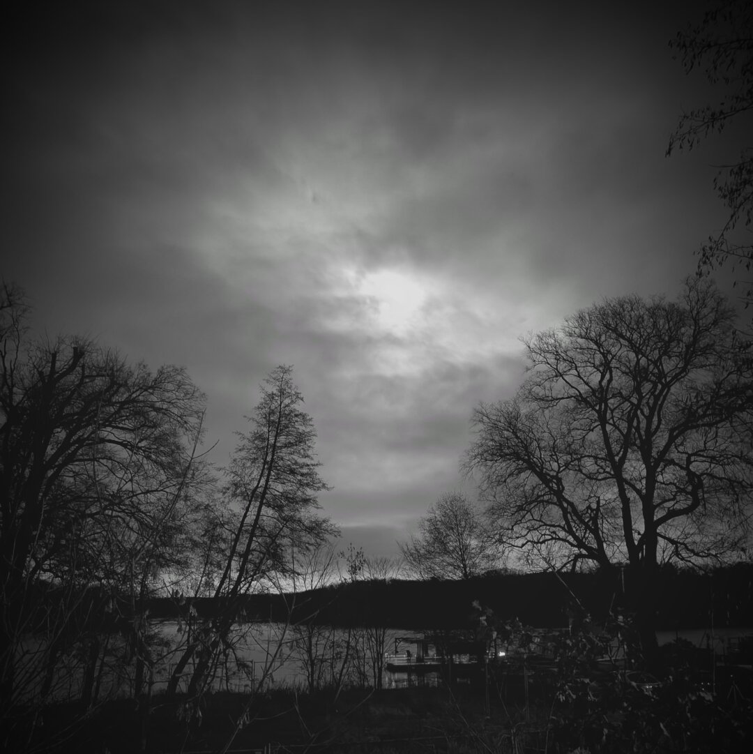 black and white photo of the moon that is behind some clouds. the landscape is lakeside with some trees