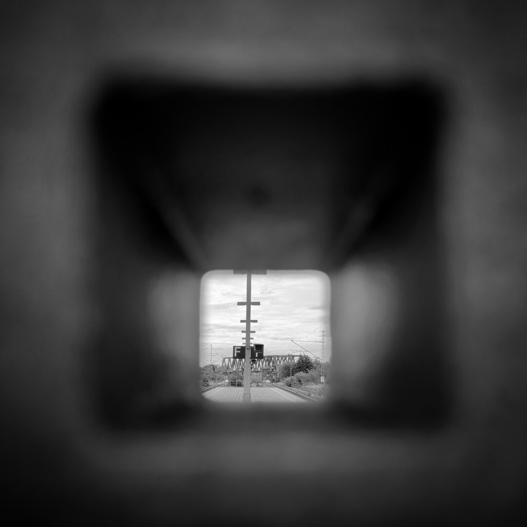 black and white photo of a train station platform. looking through a hole in a light pole.