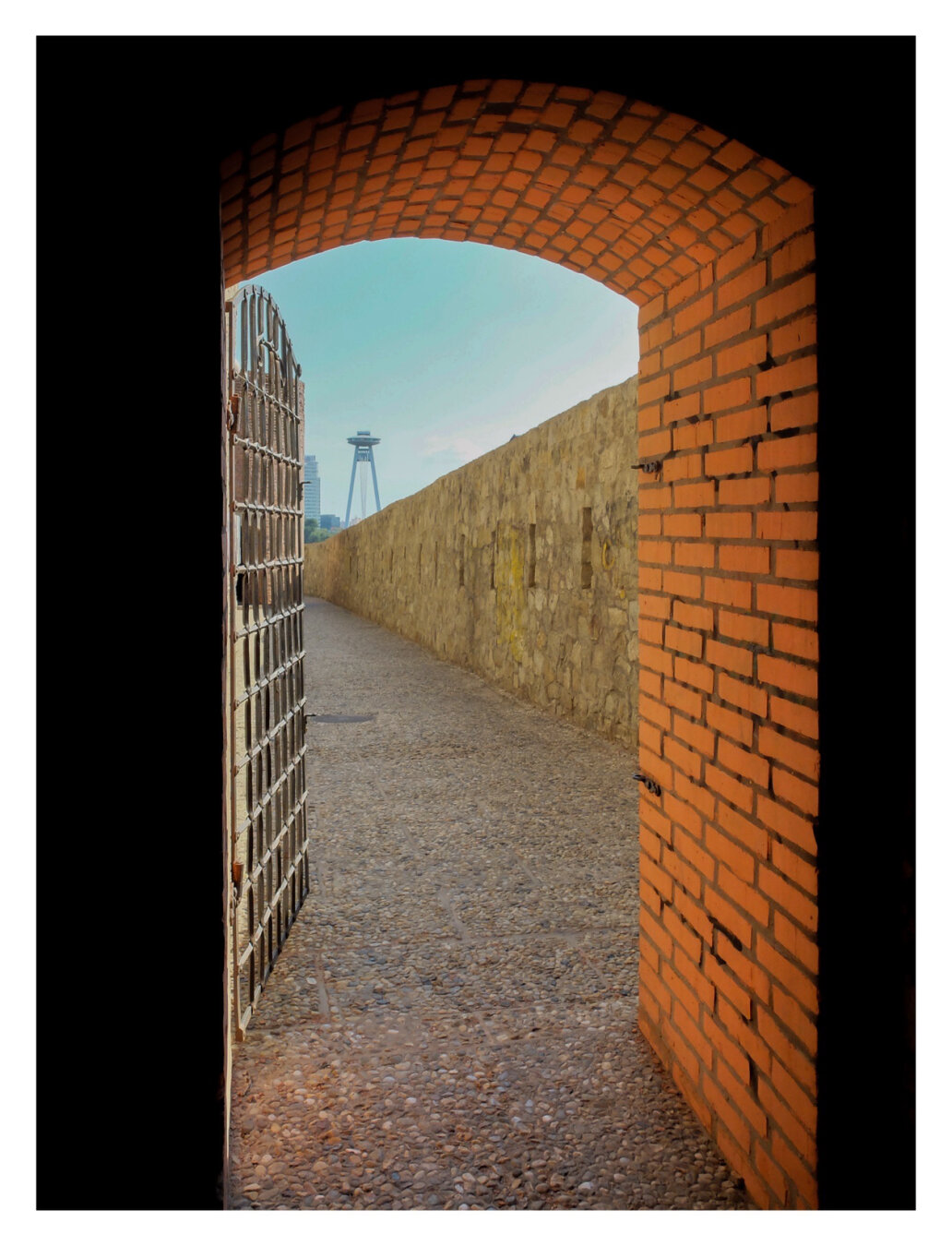 Foto im Hochformat. Blick durch einen alten Torbogen bzw.  Türbogen. Der Bogen ist aus roten Backsteinen. Alles vor dem Bogen versinkt im Schatten. Hinter dem Bogen ist eine Gittertür, die geöffnet ist, ein Weg und rechts eines Mauer. Gittertür, der obere Teil des Bogens und die Mauer Rahmen ein Stück blauen Himmel ein. Außerdem ist vor dem Himmel ein hoher Turm, der auf zwei Füßen steht und wie ein Ufo aussieht.