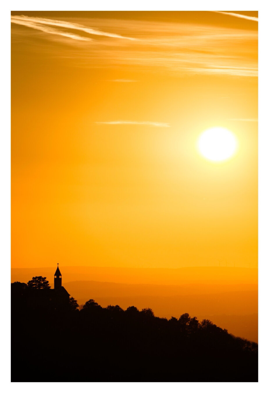 Foto im Hochformat. Kurz vor Sonnenuntergang. Unten sind die Silhouetten von Bäumen und einer Burg mit Turm. Darauf ein spitzes Dach und eine Kugel. Dahinter sind mehrere Reihen von Hügelketten, die heller werden, je weiter weg sie sind. Rechts oben ist die untergehende Sonne am Himmel, sie taucht alles in orange-goldenes Licht.