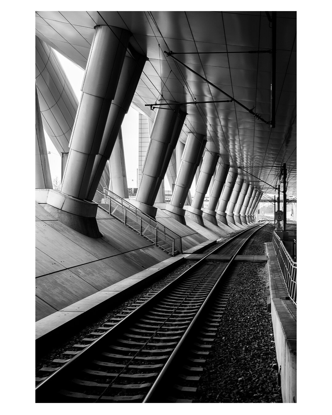 Schwarz-weiß Foto im Hochformat. Blick entlang eines Bahngleises vom Bahnhof aus. Das Gleis ist modern überdacht, leicht schräg, links vom Gleis sind in regelmäßigen Abständen dicke Säulen, die ebenfalls auf einer Schräge stehen. Das Gleis ist leer.