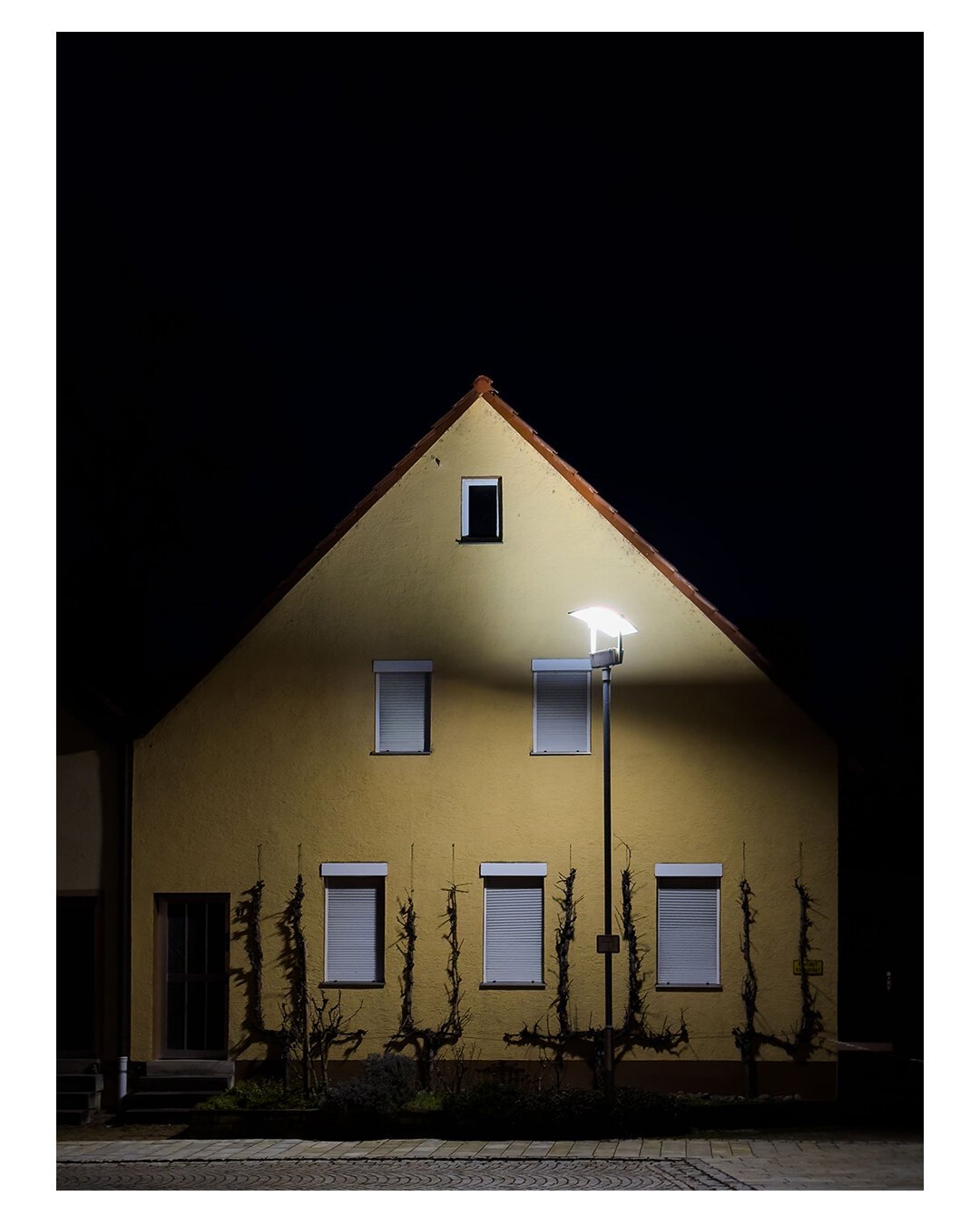 Foto im Hochformat. Es ist Nacht, Blick frontal auf ein gelbes Haus mit Spitzdach. In der Fassade sind insgesamt sechs Fenster. Unten drei, in der Mitte zwei, oben noch ein kleines. Bis auf bei dem oberen Fenster sind bei allen die Rollladen unten. In der unteren Reihe der Fenster sind dazwischen Kletterpflanzen, allerdings nur einzelne Stränge. Links ist die Haustüre. Das Haus wird von einer einzelnen Straßenlaterne beleuchtet.