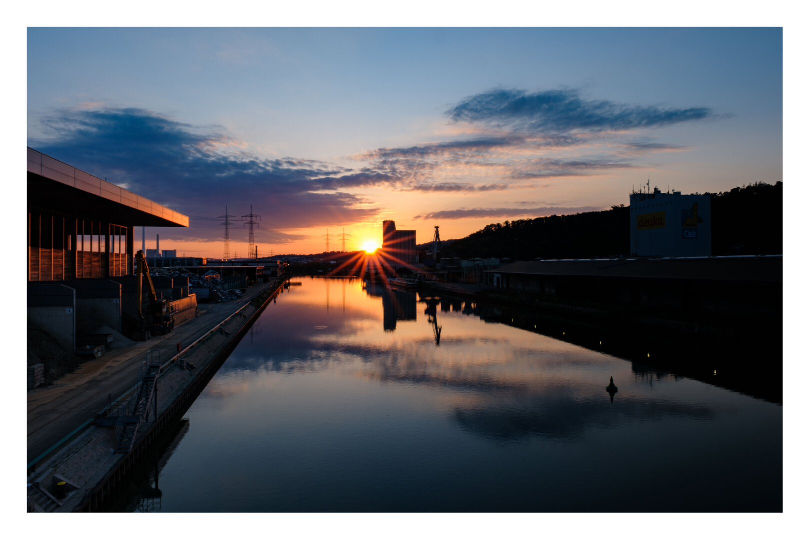 Foto im Querformat. Blick entlang eines Flusses an einem Hafen. Es ist kurz vor Sonnenuntergang. Links und rechts sind industrielle Anlagen, teils nur als Silhouette. Am Horizont ist ein höheres Gebäude, welches halb die untergehende Sonne verdeckt. Die Sonne hat Strahlen. Der leicht bewölkte orange-blaue Himmel spiegelt sich im ruhigen Wasser des Flusses.