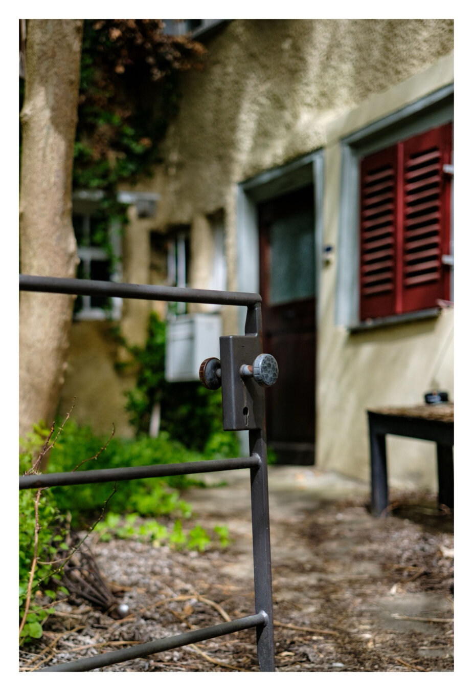 Foto im Hochformat. Im Vordergrund ist angeschnitten ein kleines Gartentörchen aus Metall. Dahinter leicht unscharf ein kleiner Weg zur Haustüre eines alten Hauses. Links und rechts sind Fenster, teils mit geschlossenen Läden, weiter links ist angeschnitten ein Baum.