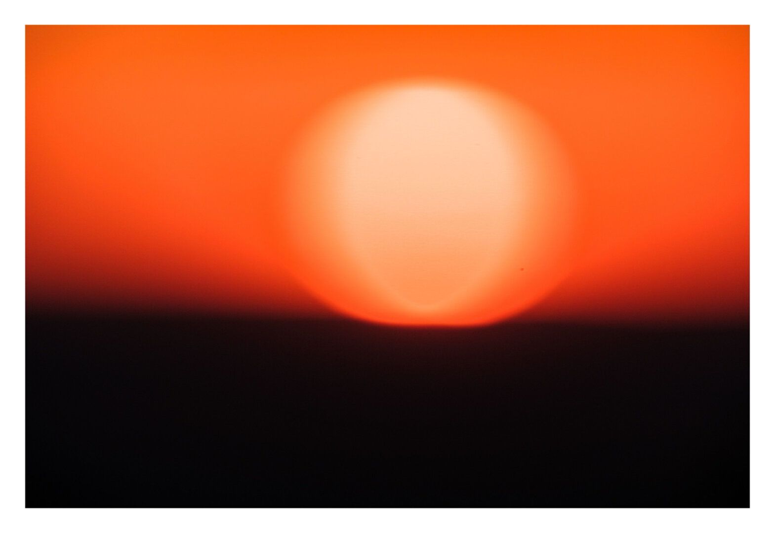 Foto im Querformat.Eine völlig unscharfe Aufnahme eines Sonnenuntergangs. Die obere Hälfte des Bildes ist rot-orangefarbener Himmel mit einer verwaschenen Sonne. Der untere Teil des Bildes ist einfach nur schwarz.