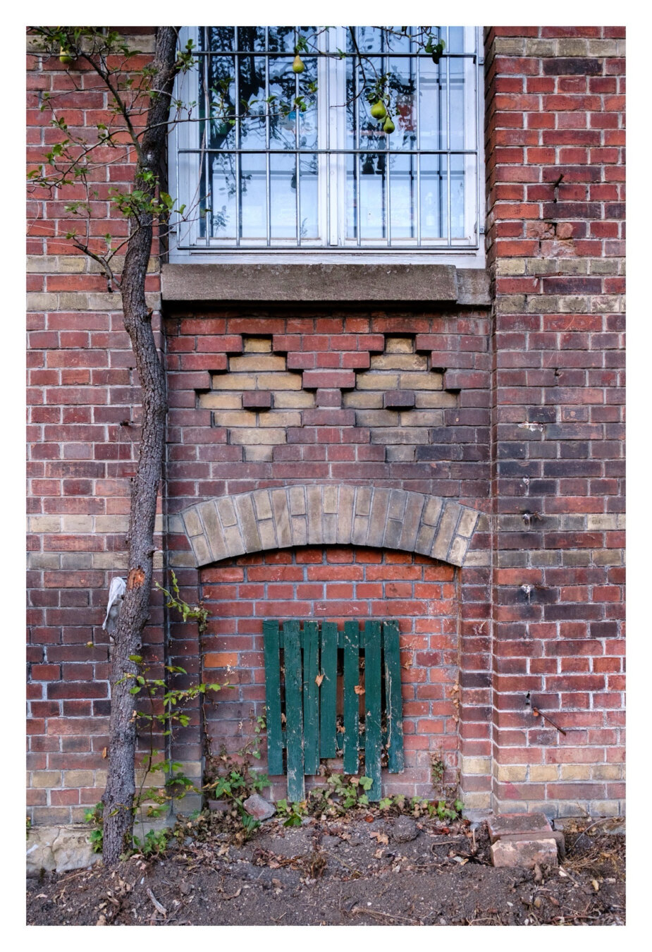 Foto im Hochformat. Eine Backsteinwand eines alten Gebäudes. Mittig oben ist angeschnitten ein Fenster mit einem Gitter davor. Unter der Fensterbank sind zwei Rauten in die Wand eingelassen. Mittig jeweils ein Stein, sehen aus wie Augen. Darunter ist ein nach oben gebogener Vorsprung, sieht aus wie ein Mund. Darunter ein weiteres Fenster, welches komplett von grünen Brettern abgedeckt ist.