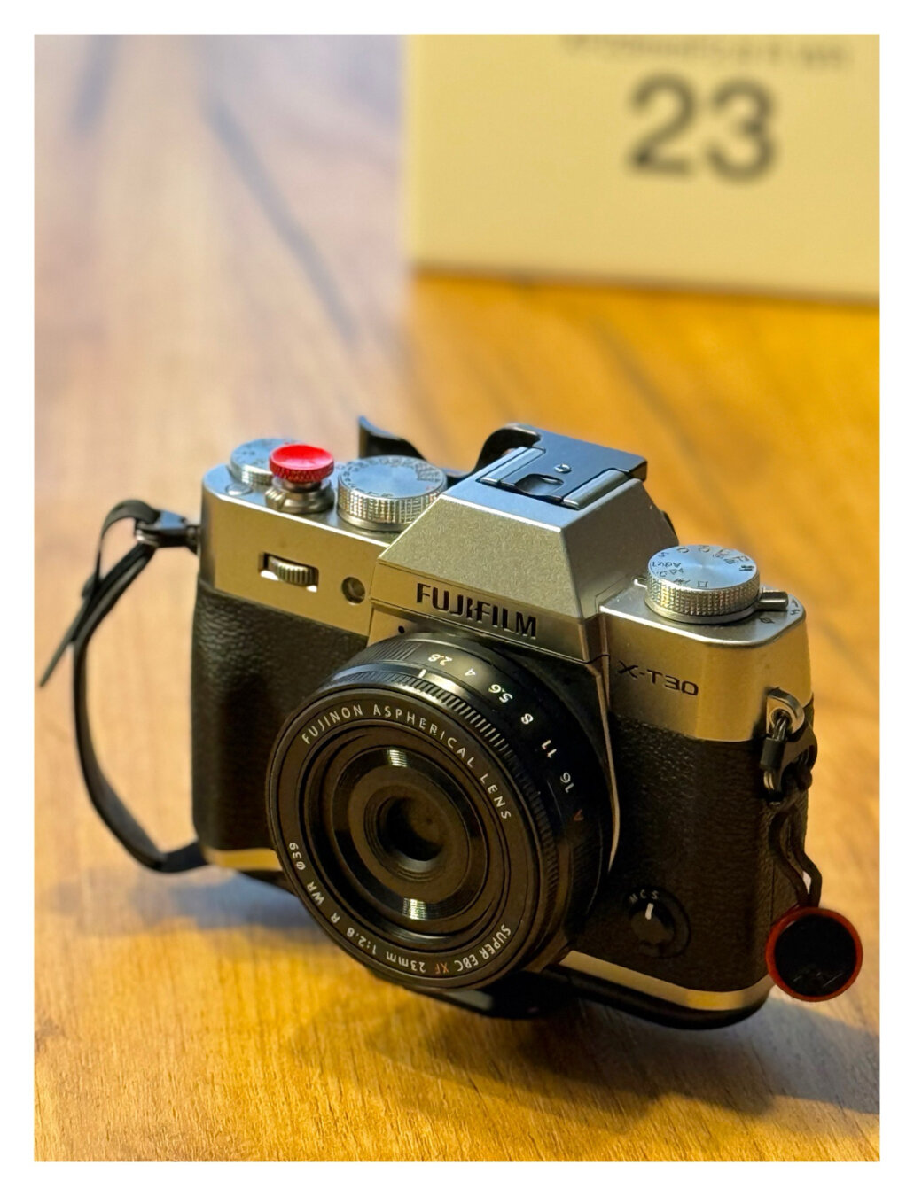 Eine Fotokamera, Fuji X-T30 II mit einem kleinen, flachen Wechselobjektiv, Fuji XF 23mm F2.8. Im Hintergrund ist angeschnitten die weiße Verpackung des Objektivs.