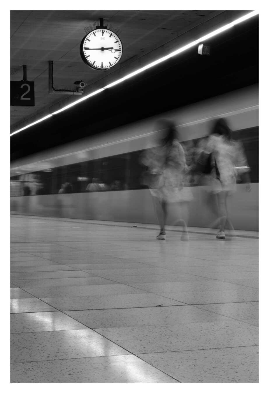 Schwarzweiß-Foto im Hochformat. Blick über ein Bahnsteig. Eine S-Bahn fährt gerade los, zwei Personen in hellen Kleidern laufen am Gleis entlang. Zug und Personen sind durch lange Belichtungszeit unscharf, die Personen fast schon Geist artig. Boden, Überdachung und eine große Uhr oben im Bild sind scharf abgebildet.