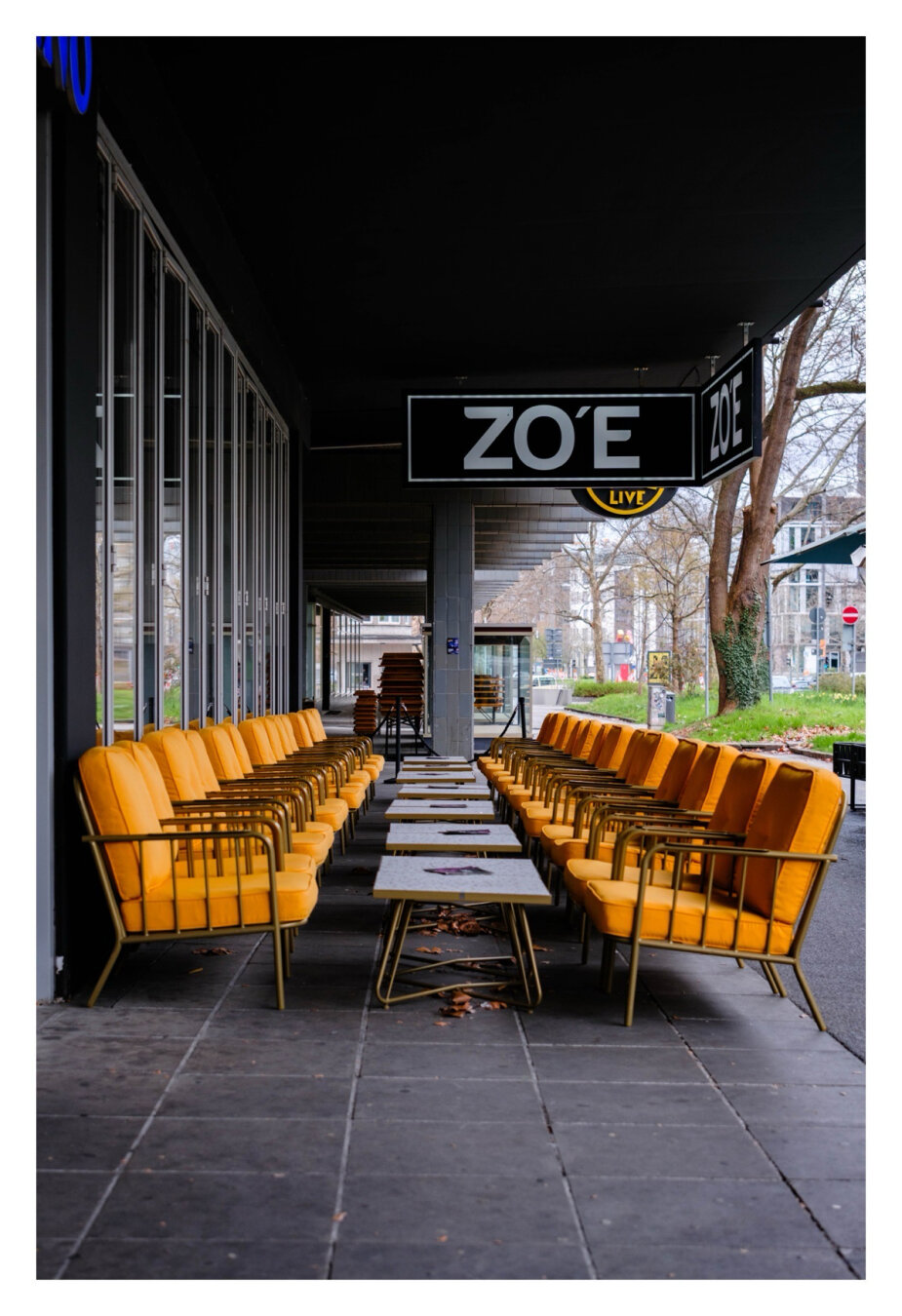 Foto im Hochformat. Der Außenbereich eines Cafés in einer Großstadt. Links und rechts sind orangefarbene Sessel aufgereiht, dazwischen jeweils ein kleiner Tisch. Auf jedem Tisch liegt eine Karte. Alles befindet sich unter einem Vordach. An diesem hängt ein Schild, darauf steht ZO‘E.