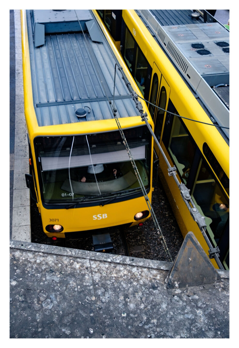 Foto im Hochformat. Blick von oben auf zwei gelbe U-Bahnen. Unten im Vordergrund ist die Kante einer dreckige Plattform, rechts ein noch dreckigeres dreieckiges gelb-schwarzes Schild, welches vor Hochspannung warnt. Die Bahnen fahren knapp nebeneinander in unterschiedliche Richtungen, die linke Richtung Kamera. Vorne ist Teil des Fahrers zu erkennen.