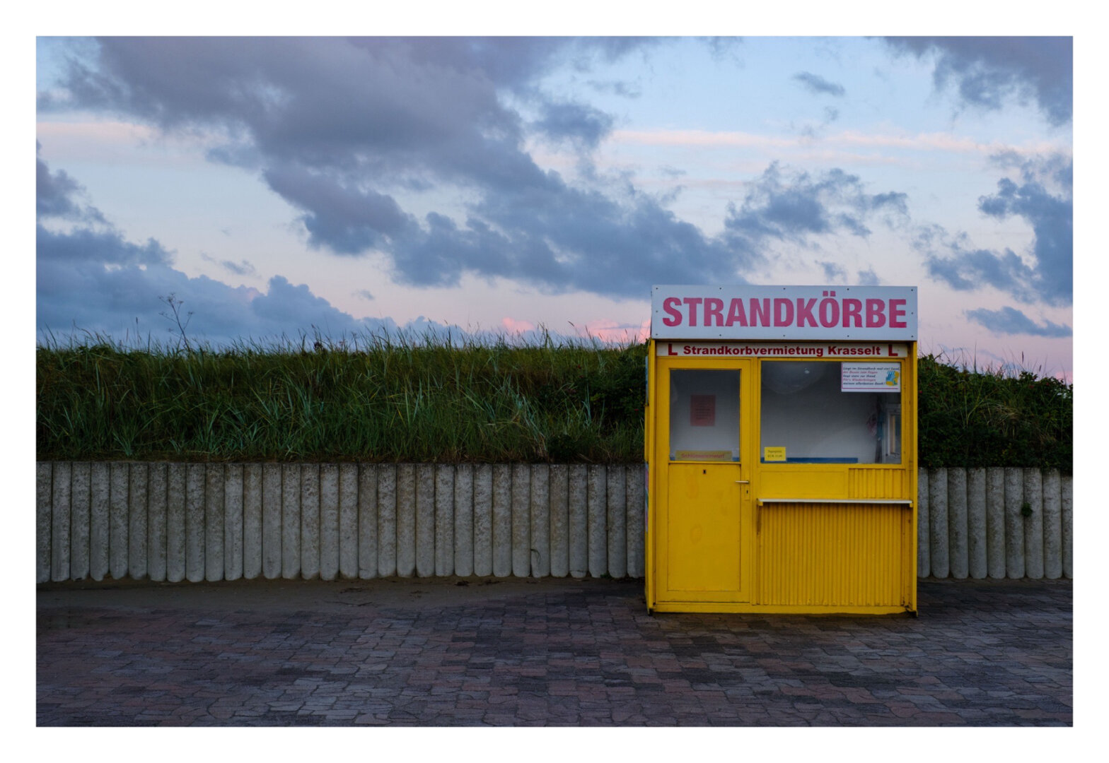 Foto im Querformat. Blick frontal auf eine kleine, gelbe Verkaufsbude. Oben ist ein weißes Schild, in roter Schrift steht Strandkörbe. Die Bude hat links eine Tür und rechts ein Fenster mit Tresen davor. Hinter der Bude ist eine ca. 1 Meter hohe Wand aus runden Steinzylindern. Darüber in etwa gleicher Höhe Gras. Die Trennlinie zwischen Mauer und Gras ist auf gleicher Höhe wie die Unterseite der Fenster in der Bude. Der Himmel darüber ist leicht rötlich und blau und mit Wolken bedeckt.