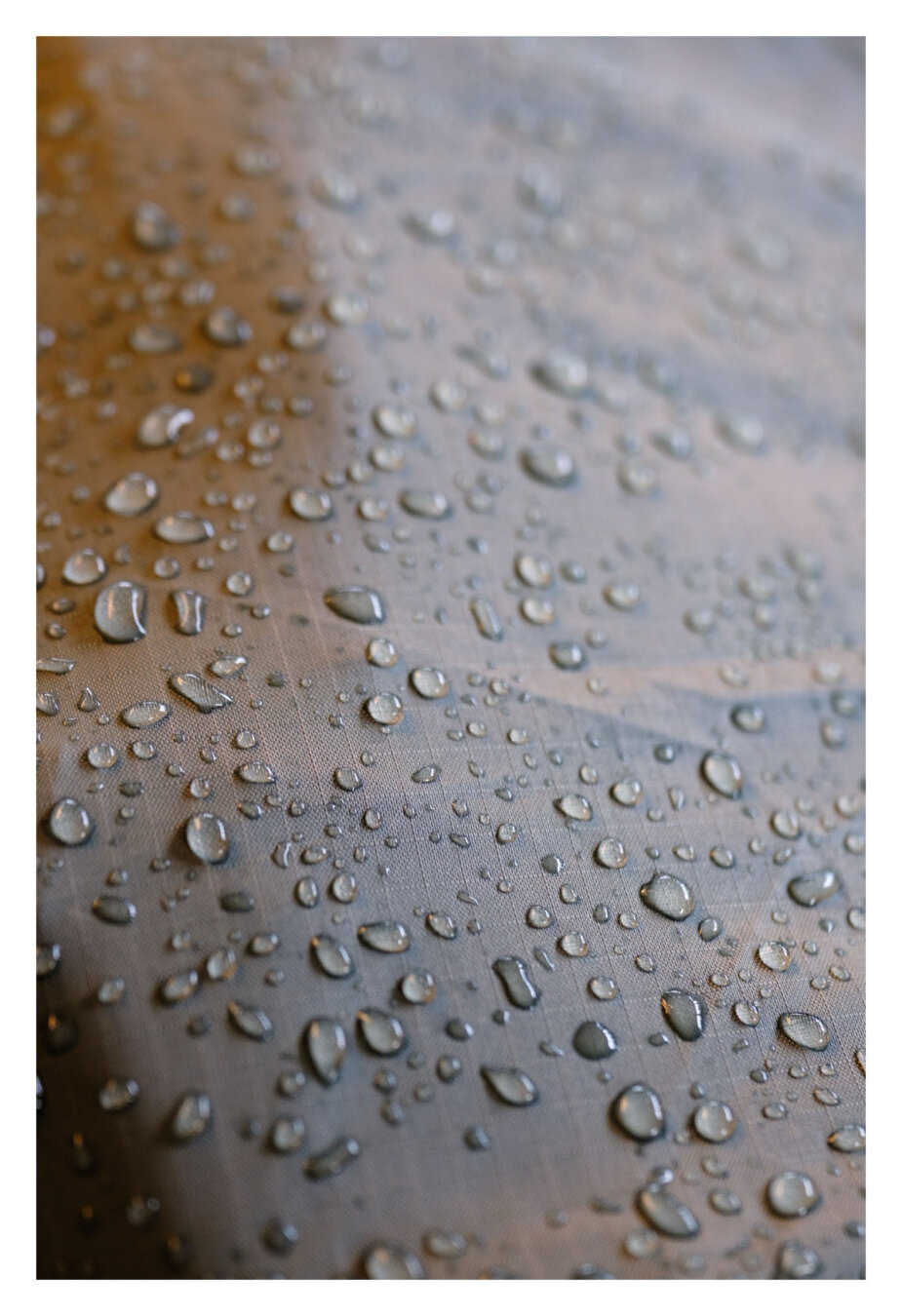 Foto im Hochformat. Nahaufnahme eines grau-bräunlichen Regenschutzes eines Rucksacks. Das Material ist sehr fein gewoben und darauf sind Dutzende von glänzenden Regentropfen.