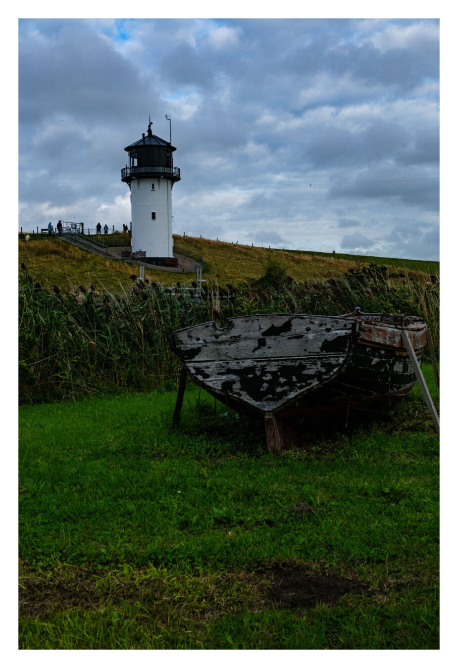 Foto im Hochformat. Blick über eine Wiese auf eine Düne. Auf der Wiese steht ein altes Ruderboot aus Holz. Weiter hinten auf der Düne ist ein weißer Leuchtturm mit schwarzer Plattform und Dach. Der Himmel ist bewölkt.