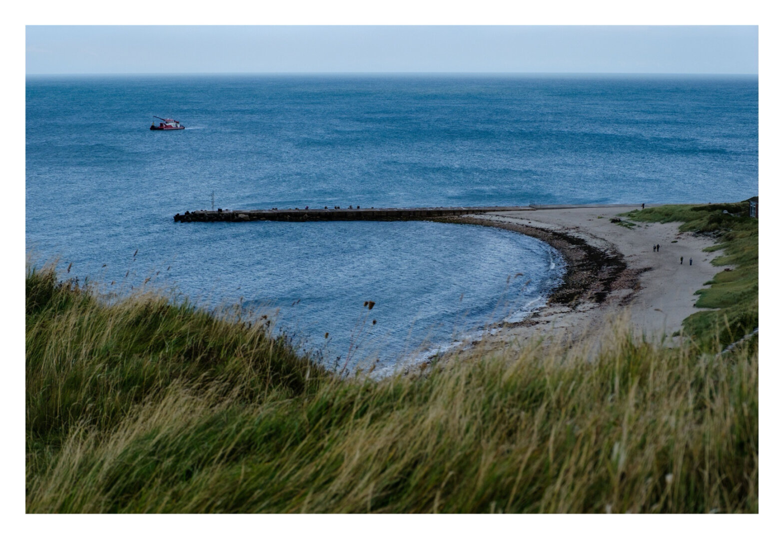 Foto im Querformat. Blick von einer Klippe auf einen sichelförmigen Strand. Im Vordergrund ist Gras auf der Klippe. Der Strand unten verläuft sichelförmig nach links und endet in einer schmalen spitze im Blauen Meer. Im Meer ist ein einzelnes kleines Schiff mit Kran darauf.