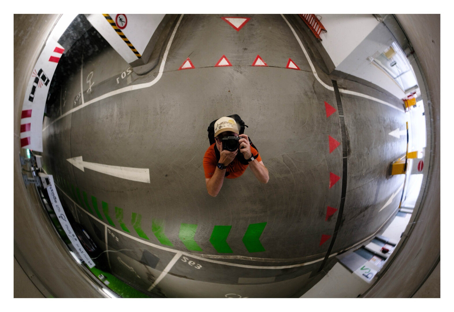 Foto im Querformat. Ich fotografier mich selbst in einem sehr weitwinkligem Fischaugenspiegel, der an der Decke in einem Parkhaus montiert ist. Ich bin dadurch mittig im Bild, orangefarbenes Shirt, helle Baseballkappe, schwarzer Rucksack, die Kamera vorm Gesicht nach oben gewandt. Um mich rum sind die grauen Wege des Parkhauses, auf denen unter anderem weiße, rote und grüne Pfeile und Vorfahrt gewähren Schilder gemalt sind.