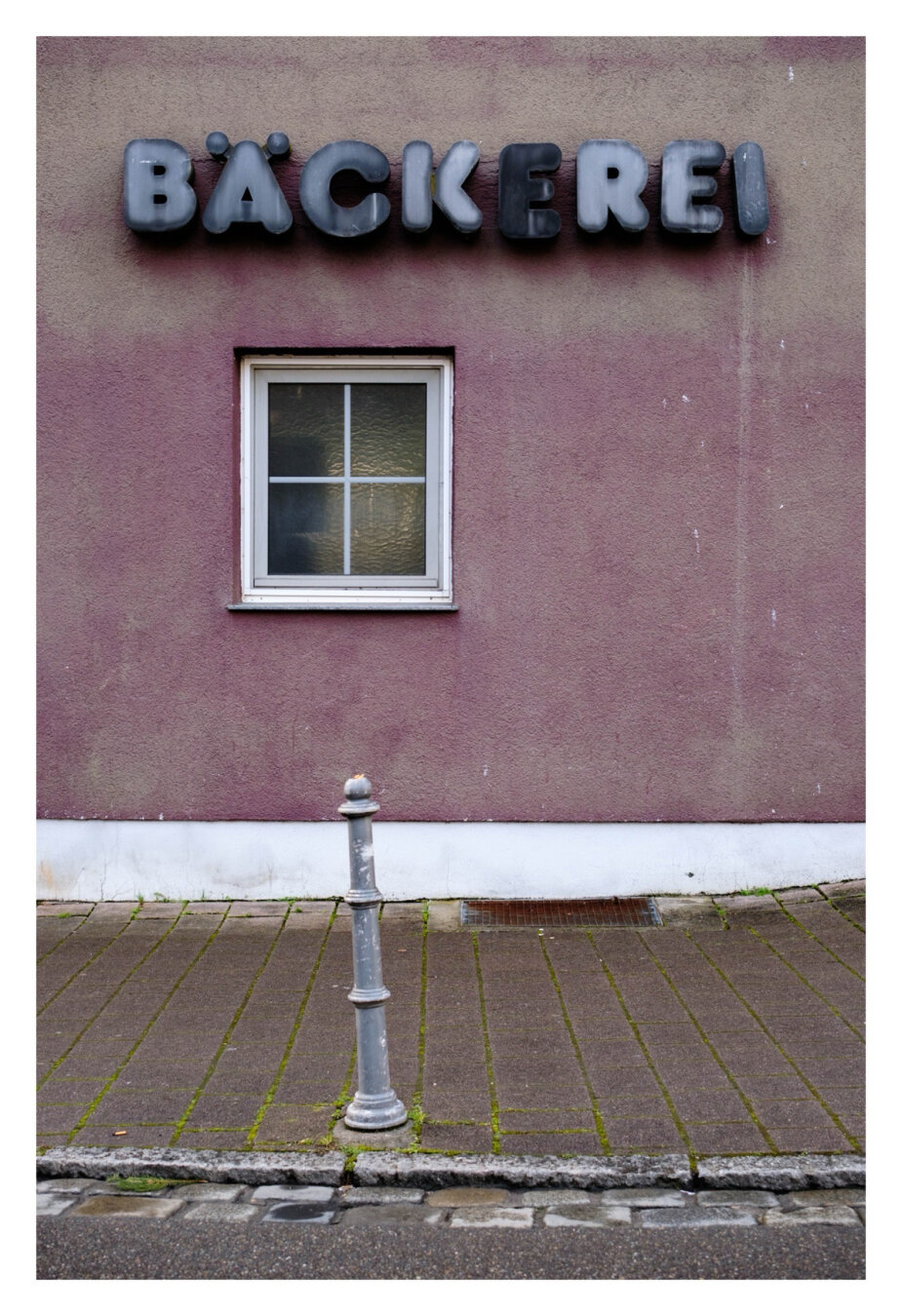Foto im Hochformat. Blick auf eine verblasste lilafarbene Hauswand. Ein einzelnes rechteckiges Fenster mit weißem Rahmen ist leicht links um Bild. Darüber steht in schwarzen verblassten Großbuchstaben Bäckerei. Unten ist an der Wand ein weißer Streifen, davor der Bürgersteig. Am Randstein zur Straße ist ein einzelner Pfosten aus Metall, im Bild ist er direkt unter dem Fenster. Der Pfosten ist leicht schräg.