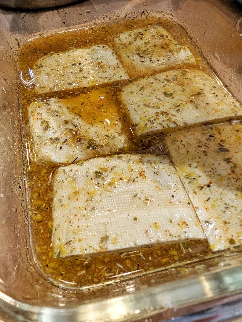 Eckige Glasschale mit ölig-wässriger Flüssigkeit voller Gewürze.  In dieser Marinade ruhen mehrre große, weißliche Stücke Tofu