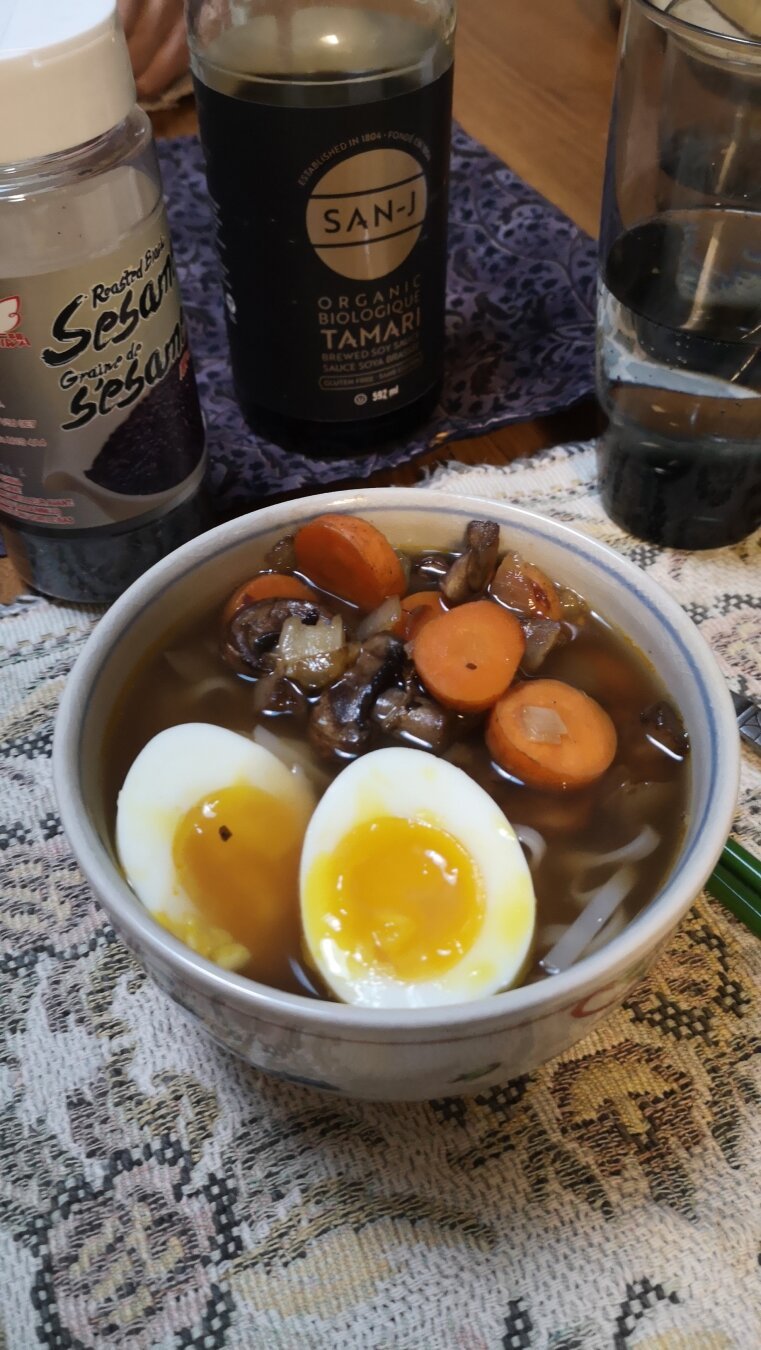 Picture of a bowl with a brown broth, rice noodles, a halved, soft-boiled egg, and fried carrots, onions, and mushrooms. There is a bottle of tamari sauce and a shaker of sesame seeds in the background.