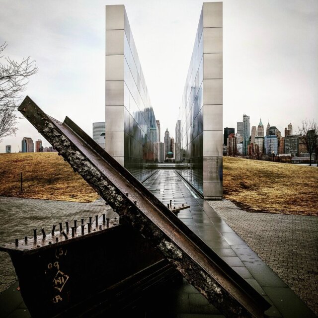 Monumento minimalista de dos muros de acero pulido entre los que se recorta una ciudad de rascacielos al fondo. En primer plano, una viga retorcida del derrumbe original, oxidada y conservada. La memoria también pide arquitectura.