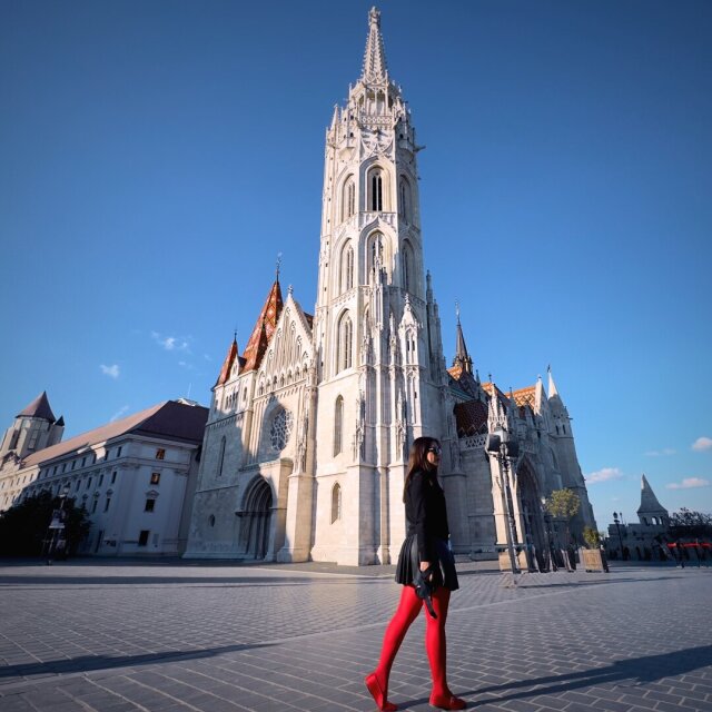 Mujer caminando de perfil con medias rojas, vestido y chaqueta negros, frente a una iglesia gótica blanca con aguja afilada y tejado de tejas vidriadas de colores. La plaza vacía la deja sola con la fachada. El rojo eligió aparecer donde nada más lo iba a hacer.