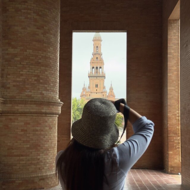De espaldas, una mujer con sombrero de paja de ala ancha y melena larga fotografía un torreón de ladrillo visto a través de un vano arquitectónico. Las dos columnas macizas la enmarcan como visor de la propia escena. Toda foto incluye al fotógrafo aunque pretenda lo contrario.