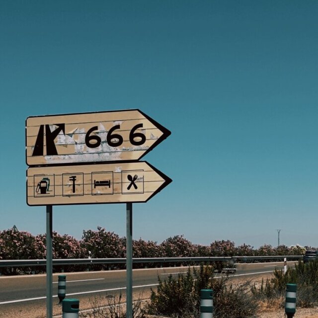 Señal de salida de autopista con el número 666 sobre indicador de gasolinera, alojamiento y restaurante. Cielo azul saturado, adelfas y balizas blancas y verdes en la cuneta. La carretera elige sus propios símbolos sin pedirle permiso a nadie.