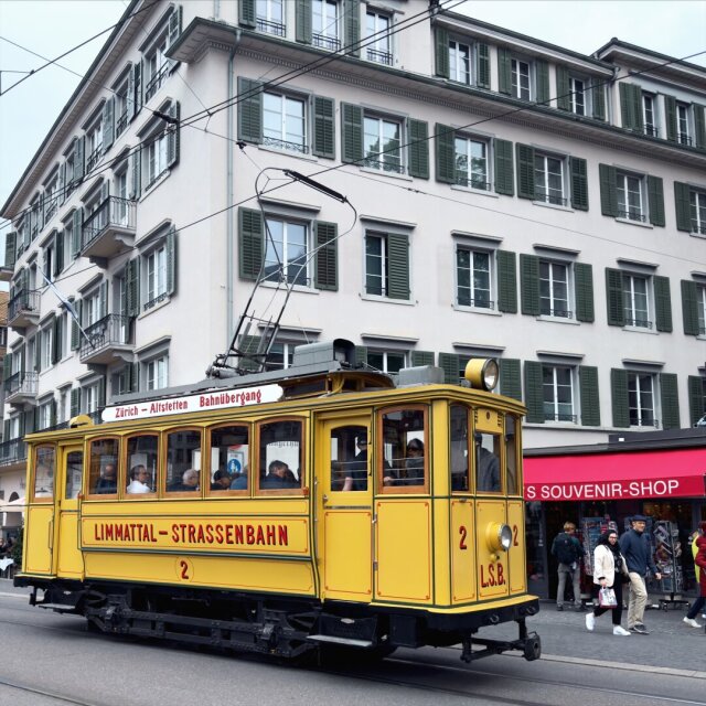 Un tranvía amarillo de otra época, Limmattal-Strassenbahn nº2, parado contra una fachada de persianas verdes y balcones de hierro. Pantógrafo en alto, faro como ojo somnoliento, los pasajeros asomados como si posaran sin saberlo. A la derecha, una tienda de souvenirs en rojo brillante les hace coro. El pasado vuelve a la calle a repartir parada y, durante un trayecto, todo el mundo finge que no ha pasado el tiempo.